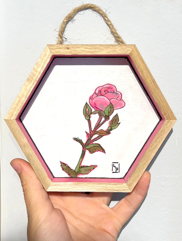 A hexagonal wooden shadow box hanging display held in a hand, featuring a painted pink rose on a white background with a pink inner border.